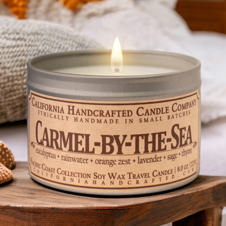 A lit soy wax travel candle from California Handcrafted Candle Company, labeled ‘Big Sur,’ is displayed on a wooden table with a blurred ocean view in the background. The label reads ‘Ethically handmade in small batches’ and features scent notes of golden sage, beach juniper, sea salt, and island gardenia. The candle is part of the Pacific Coast Collection and is housed in an 8 oz. silver tin.